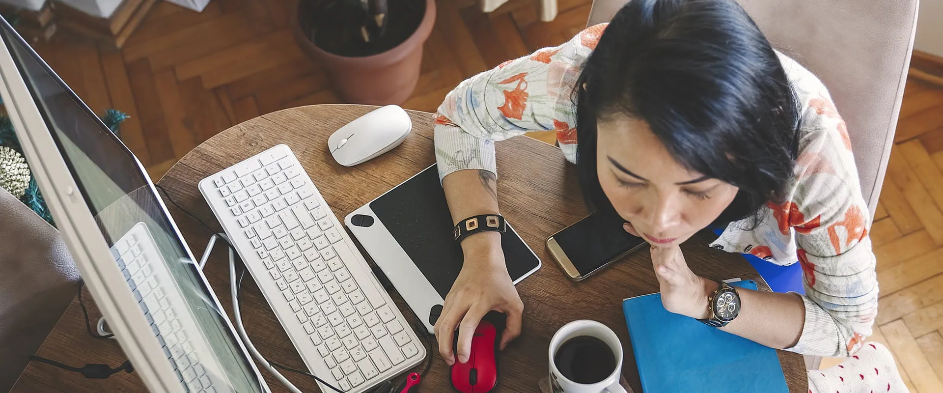 An individual sits at their desk with a desktop computer, laptop, phone, notebook, and cup of coffee, during their job search.