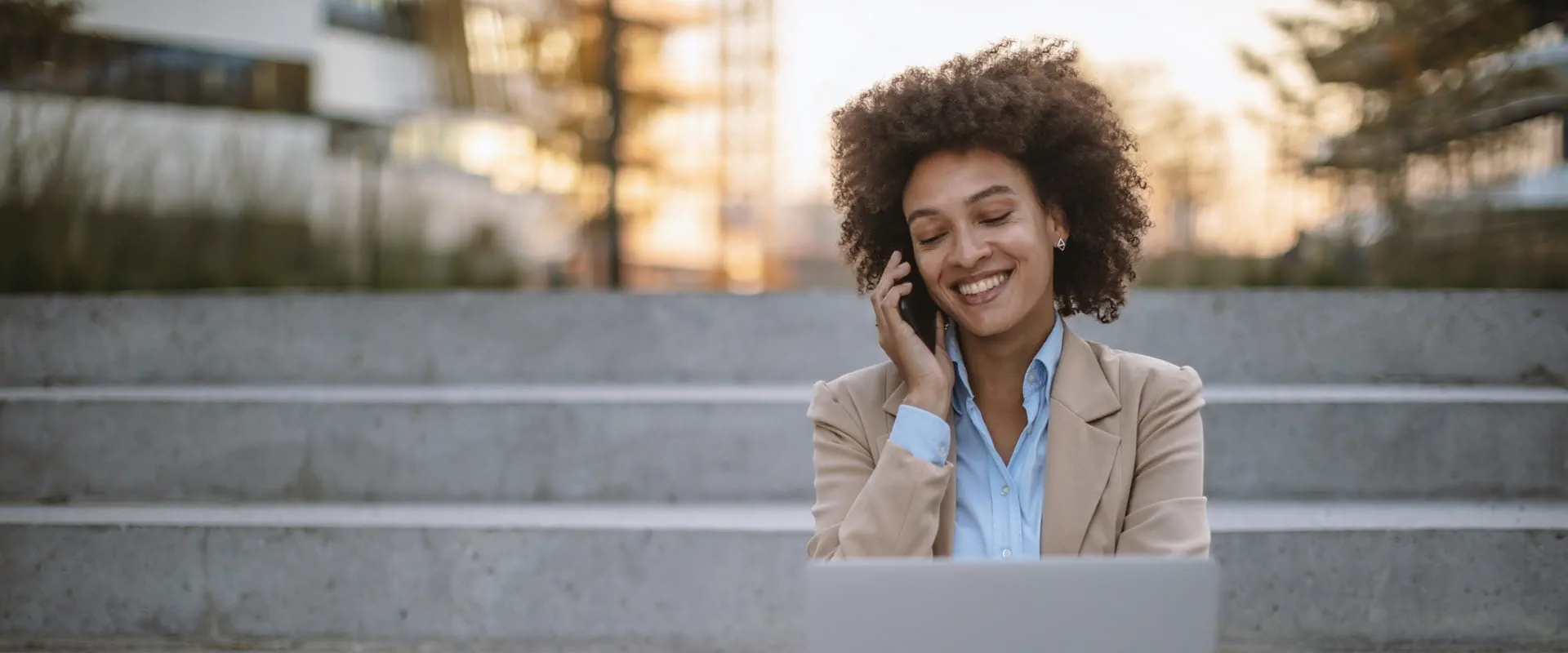 Person sitting outdoors on steps, engaged in a phone conversation while working on a laptop, smiling in a professional setting.