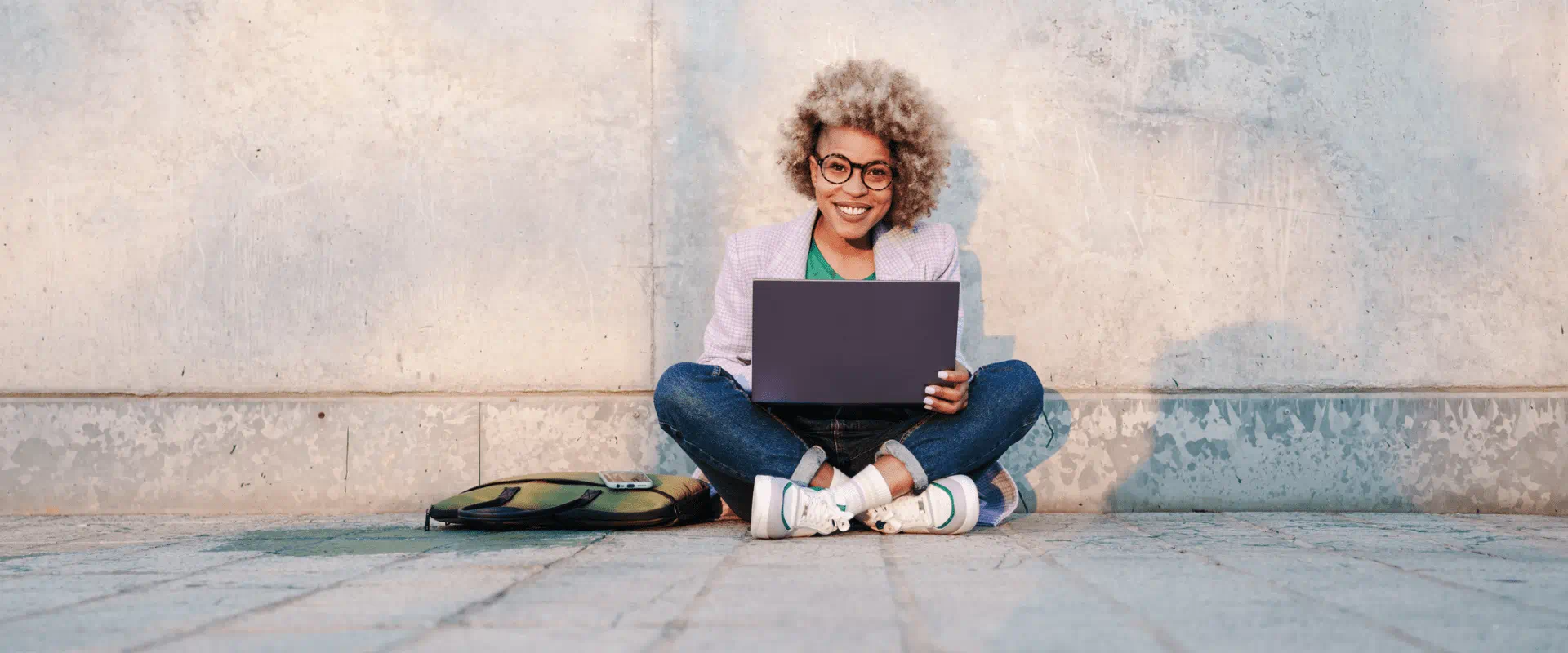 A smiling person sits outside with a laptop in their lap.