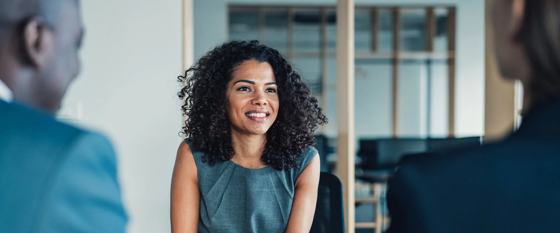 A confident professional sitting in a meeting room, engaging with colleagues who are partially visible.