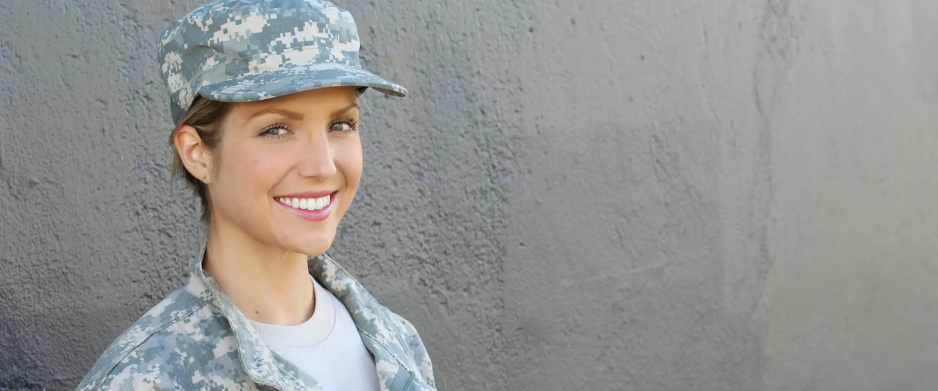 Person in military uniform smiling while standing against a textured wall.