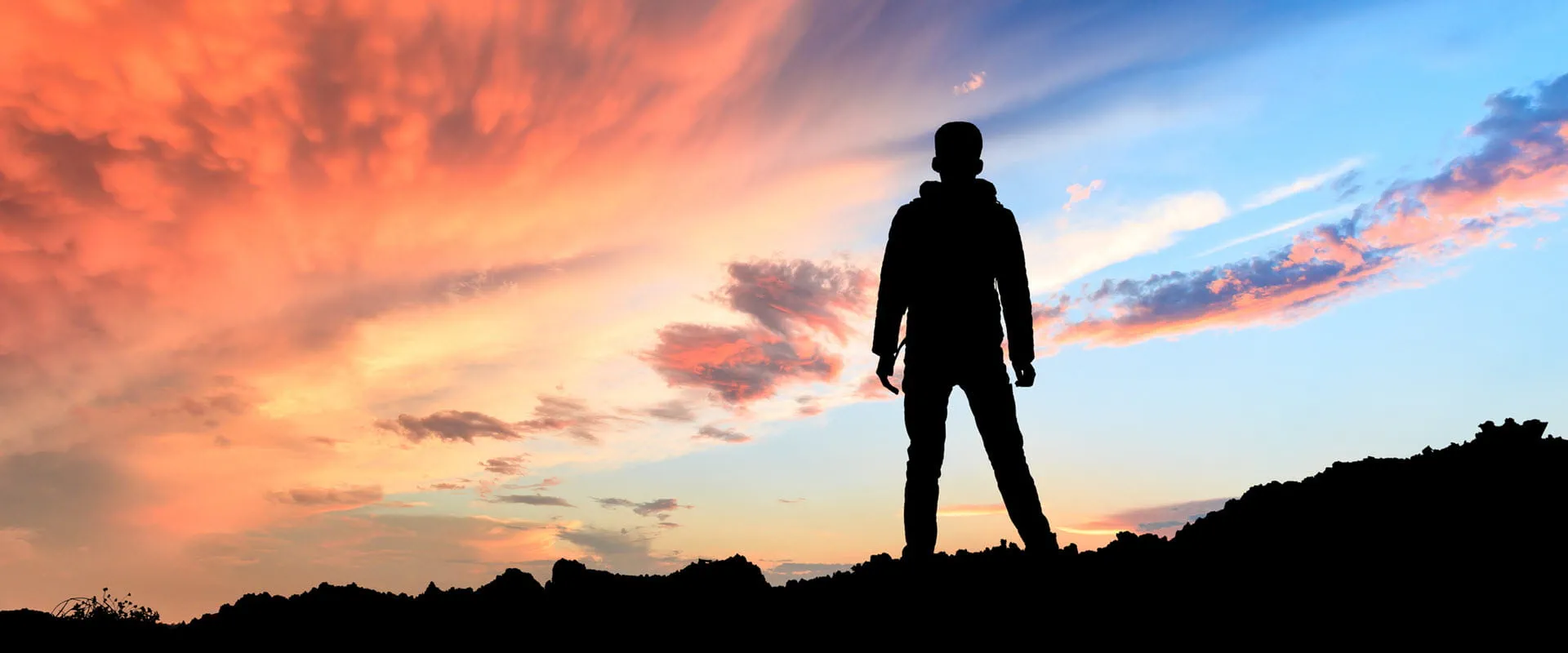 Silhouette of a person standing on a hilltop against a dramatic sunset sky with vibrant orange, pink, and blue hues.