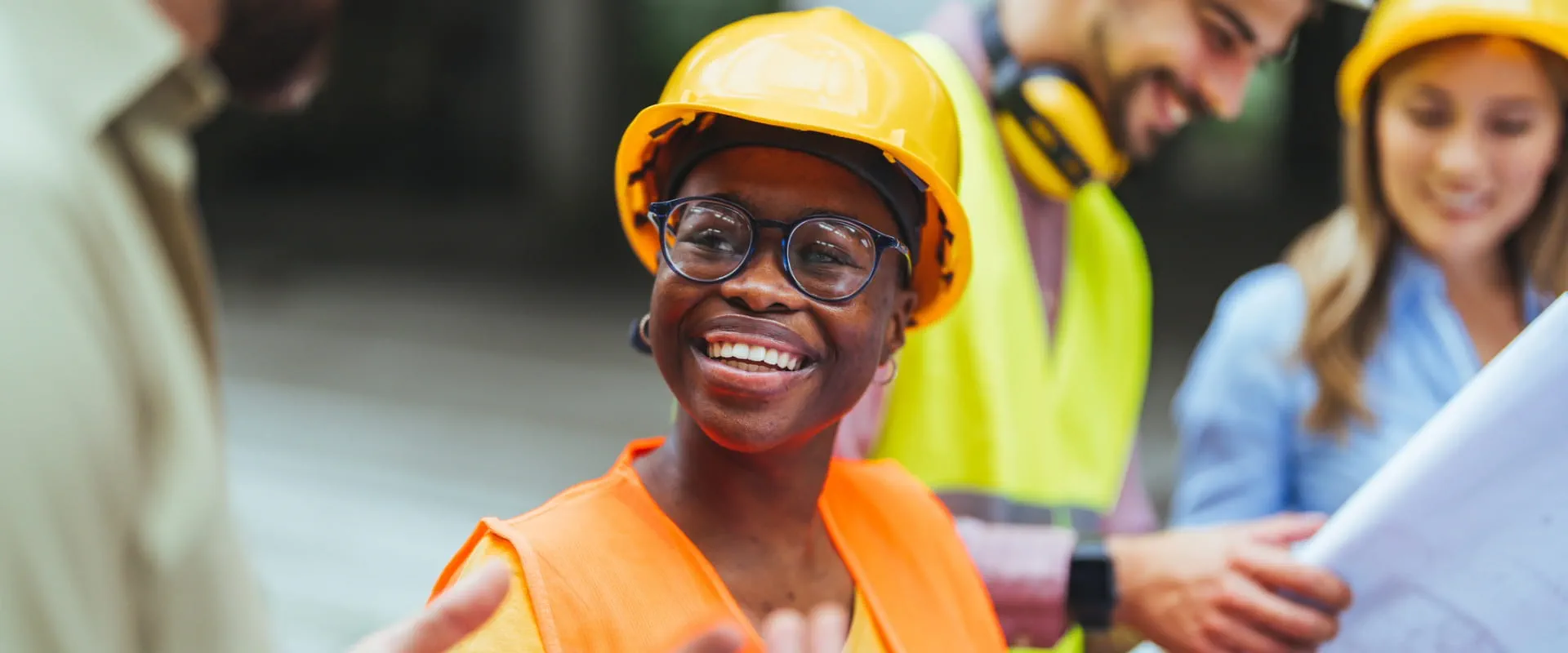 Worker in a hardhat and safety vest stands in a crowd of similarly dressed workers, smiling.