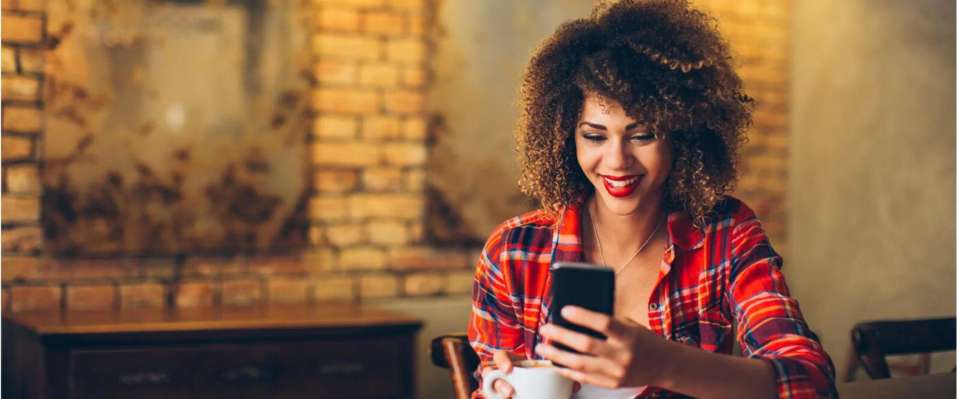 Smiling person in a coffee shop, wearing a red plaid shirt, enjoying a cup of coffee while looking at her smartphone.