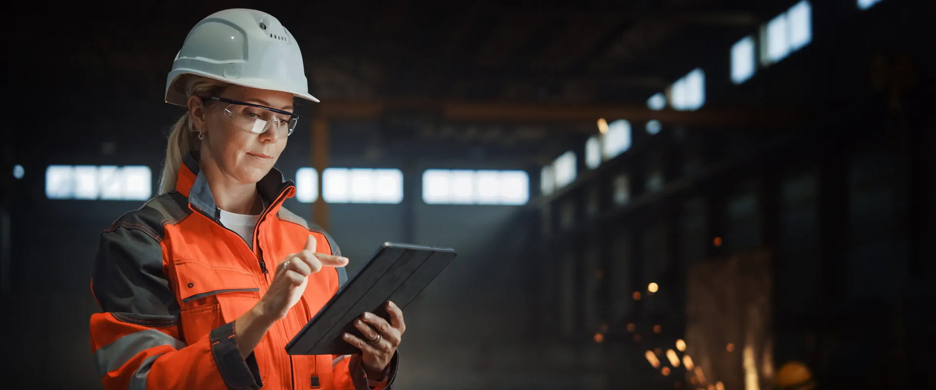 Worker in a hard hat and protective eyewear uses a tablet in an industrial setting,