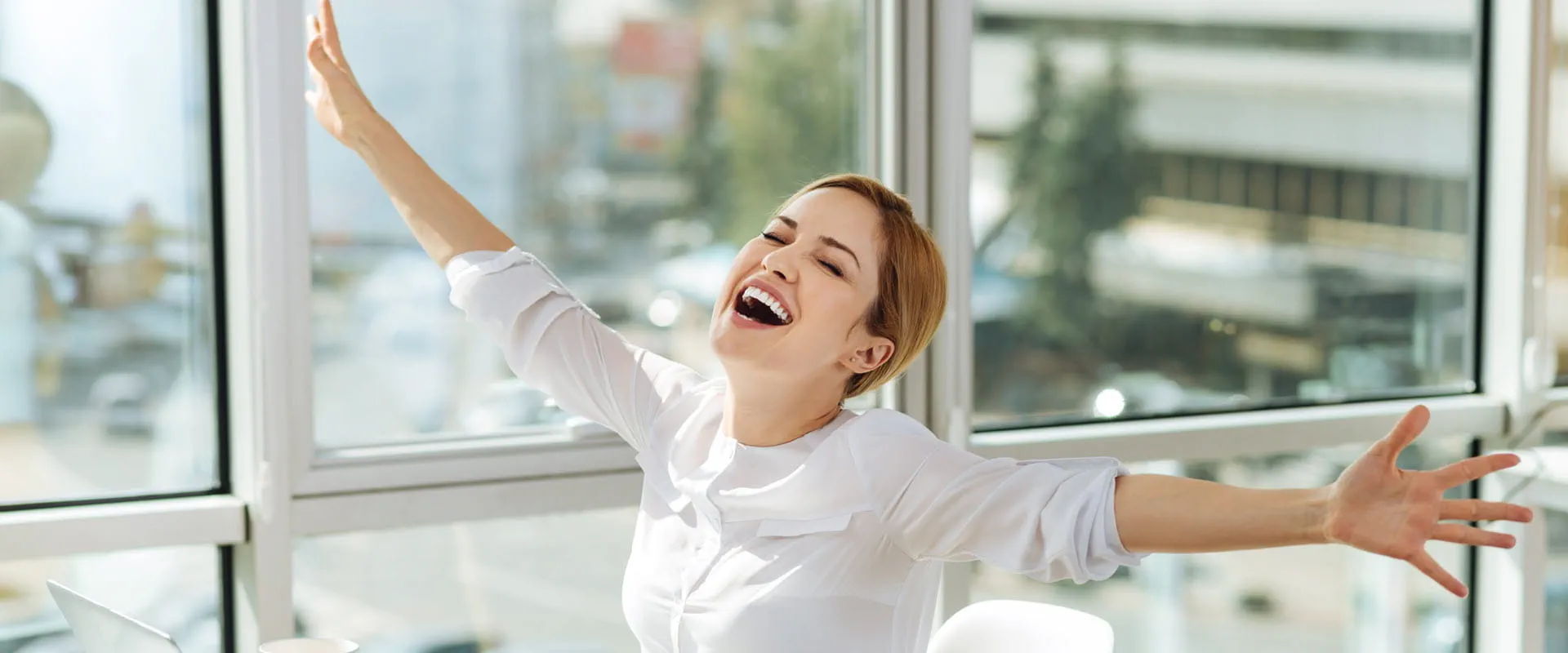 Excited person in a bright office, smiling with arms outstretched, celebrating a successful moment.