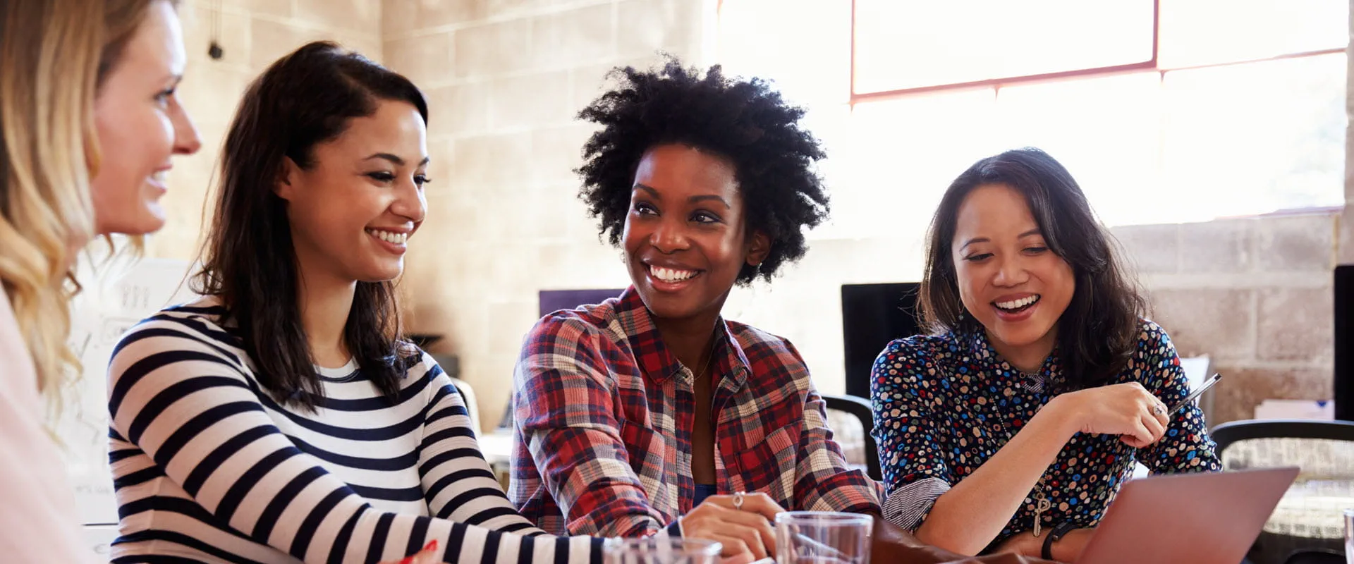 Four people sitting at a table, smiling and collaborating while using a laptop in a casual office setting.