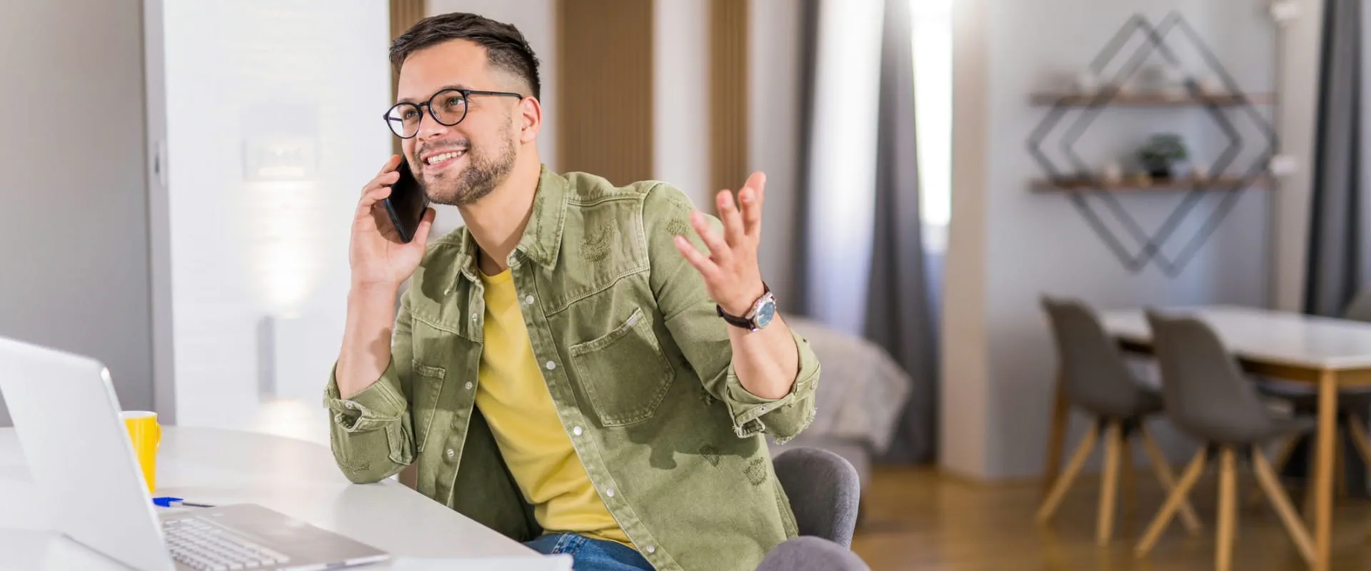A casually dressed professional having a phone call while working from home, sitting at a desk with a laptop