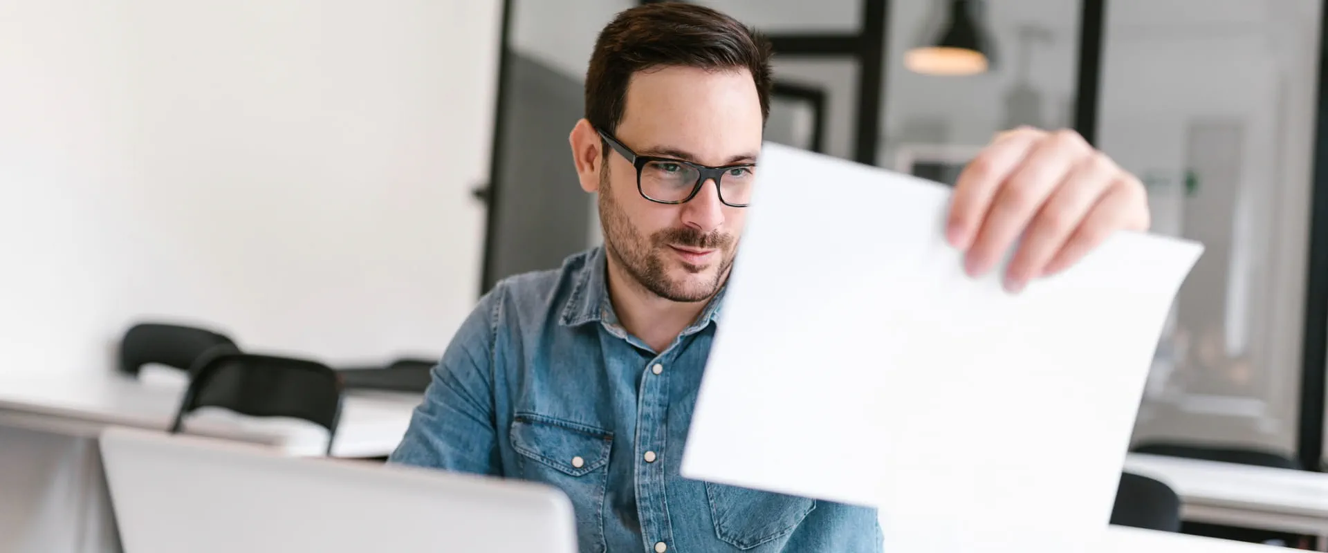 Sitting at a desk with a laptop, an individual looks closely at a piece of paper in their hand.