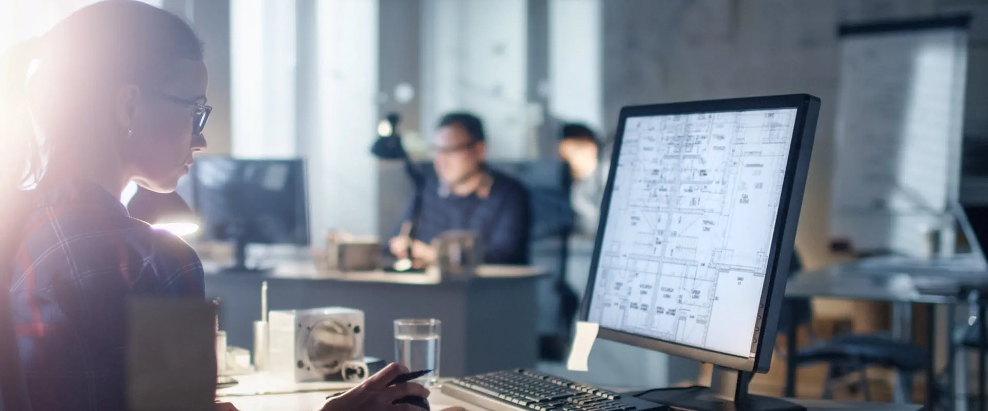 A person working at a desk in a dimly lit office, reviewing blueprints on a computer monitor, with colleagues working in the background.