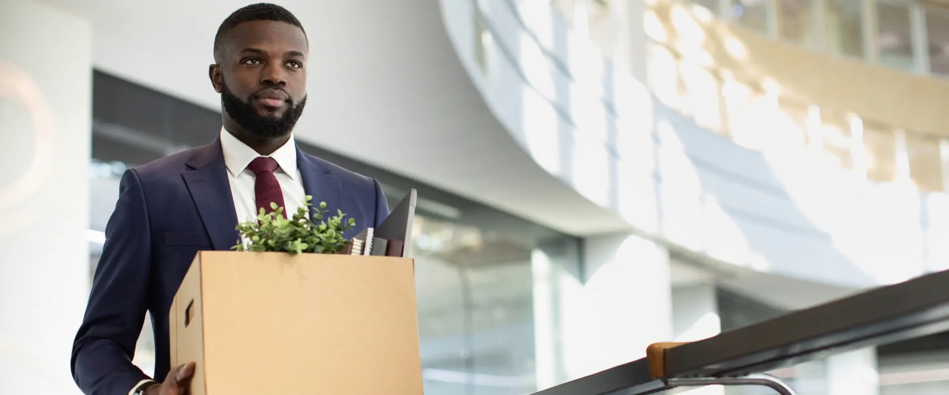 A worker in a suit seriously walks through the lobby of a large office building. He is carrying a box filled with personal items.