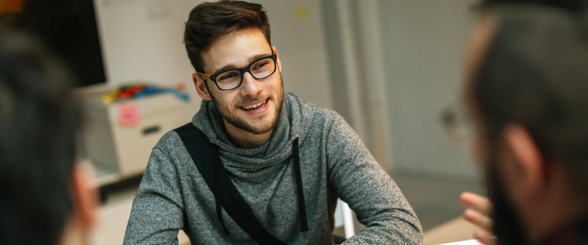 A young person with glasses, casually dressed, leans forward on a desk listening to someone.
