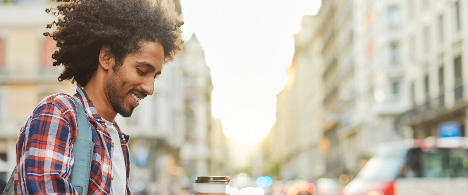 A smiling individual walks down a city street, looking at his phone and holding a cup of coffee.