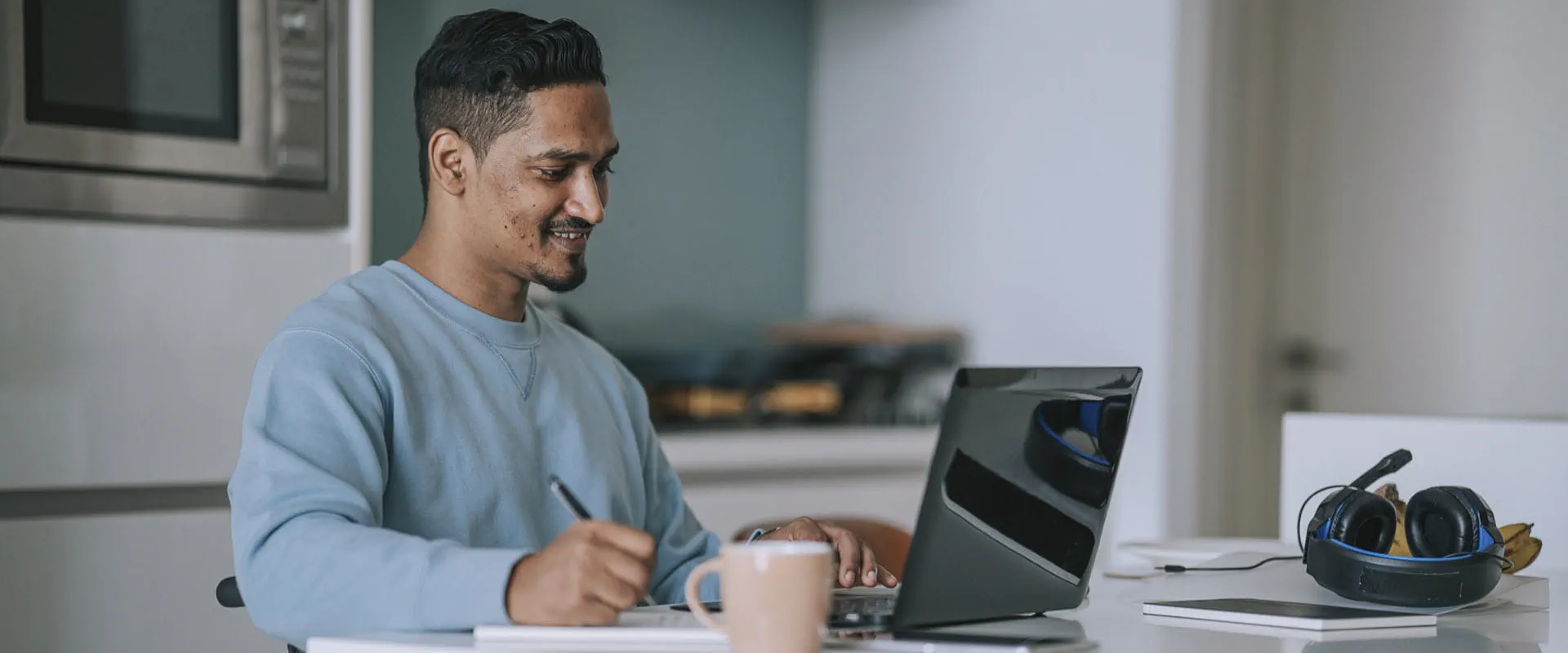 A person in a wheelchair is at their kitchen table, writing something in a notepad while looking at their laptop.