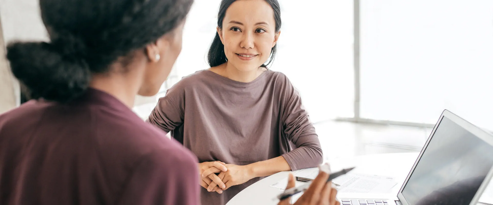 Two people sitting at a desk having a professional conversation, with a laptop open in front of them.