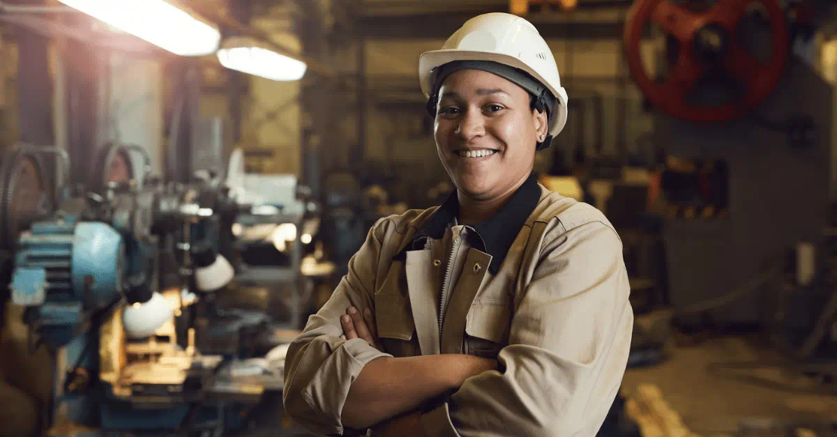 A worker in a hard hat and coveralls smiles at the camera. They are in a manufacturing floor.