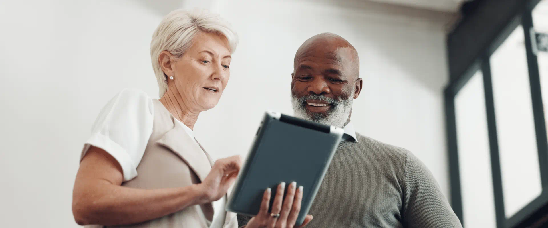 Two older workers stand in an office looking at a tablet.