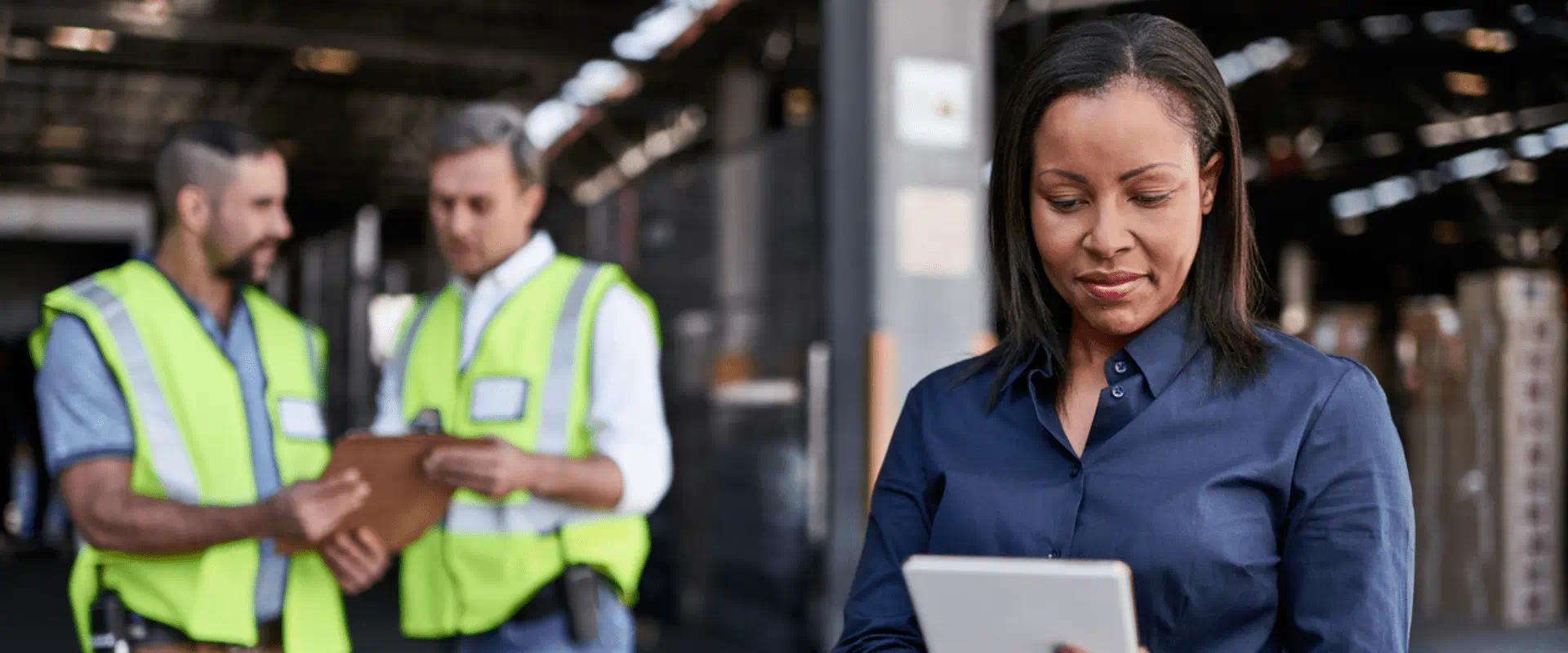 A worker looks at a tablet, while two others in safety vests talk behind them.