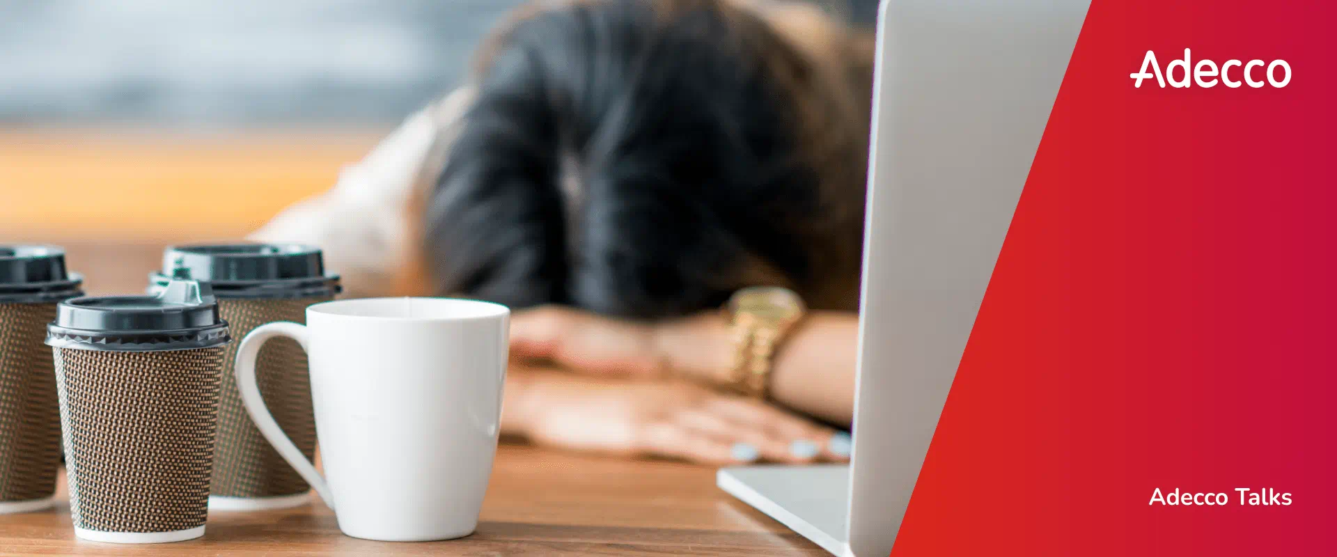 A worker sits with their head laying on their desk. There are three coffee cups and a laptop on the desk.