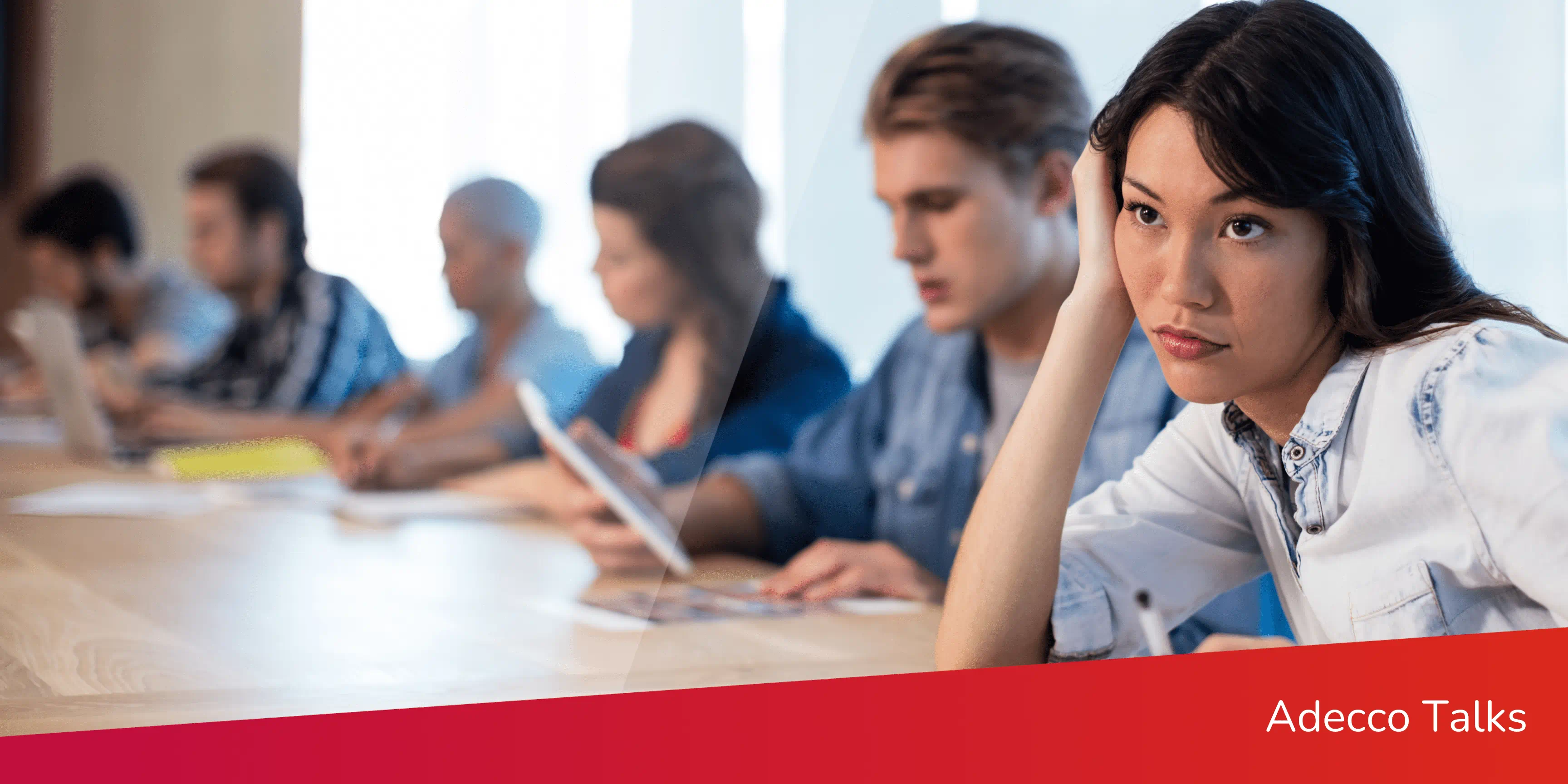 A worker sits at a conference table with coworkers. They look bored and annoyed