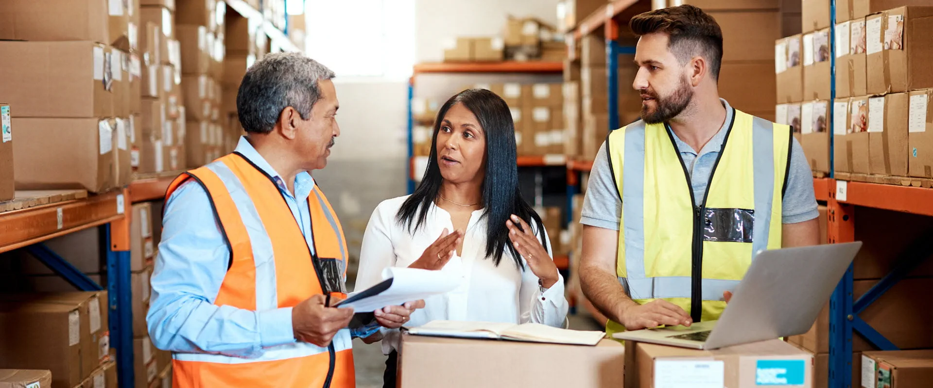 Three logistics workers, two in safety vests, have a meeting on the warehouse floor.