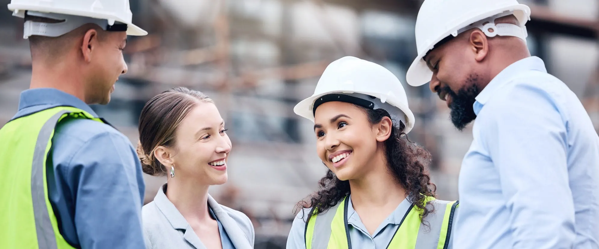 Four workers in a combination of hard hats and business clothes stand together and talk about work.