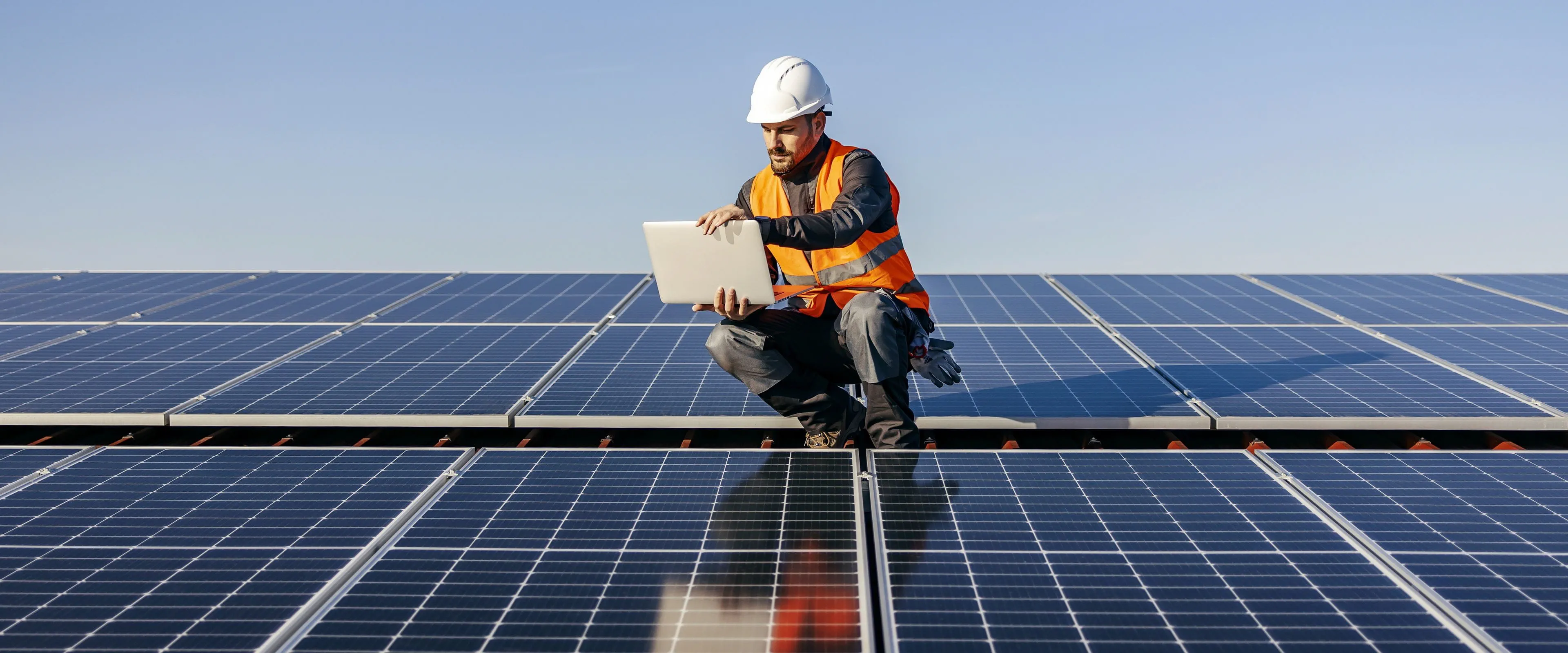 A worker in a safety vest and hard hat looks at a tablet, while crouching on a roof covered in solar panels.