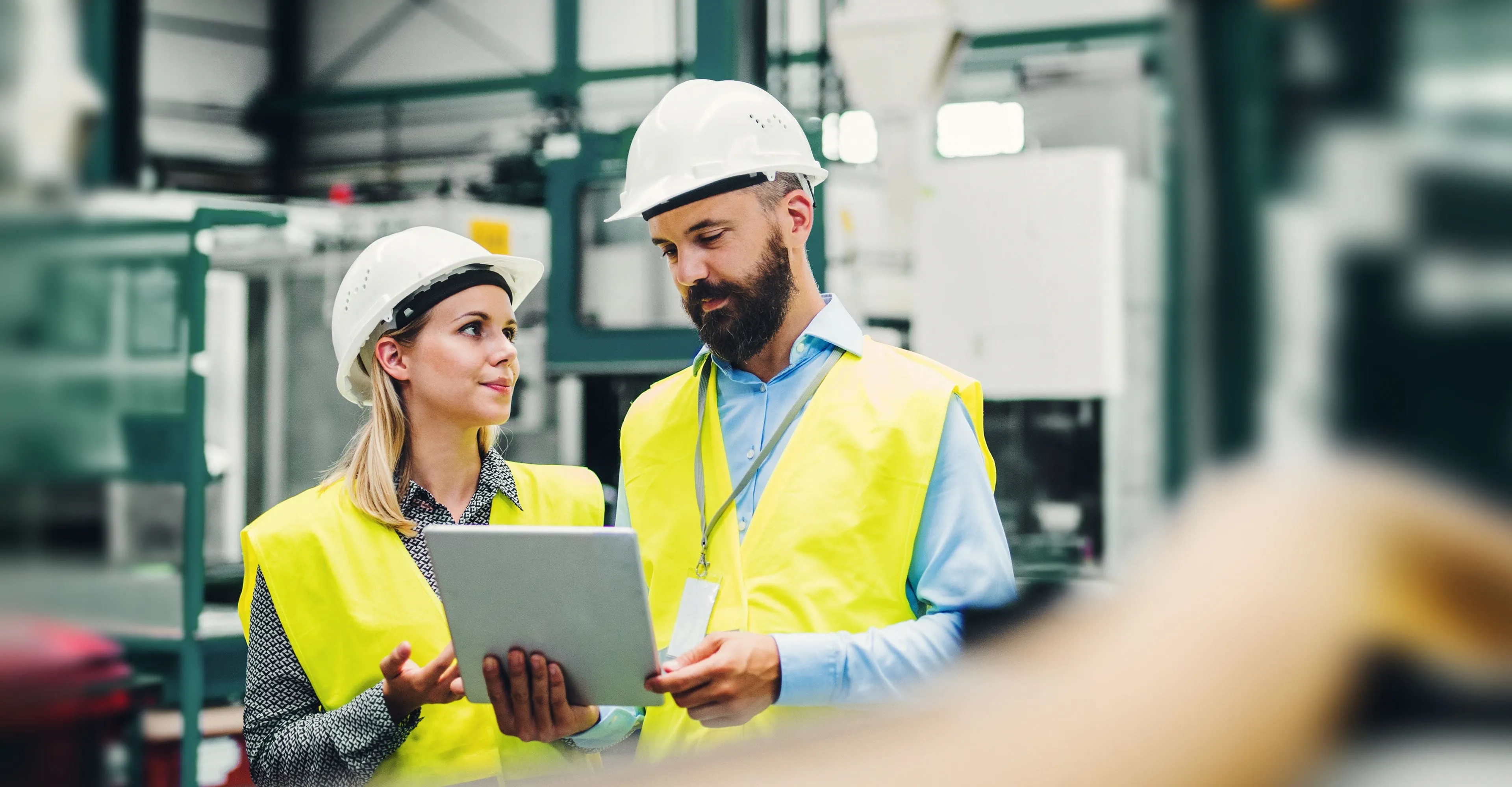 Two factor workers in business casual clothes, safety vests, and hard hats look at a tablet.