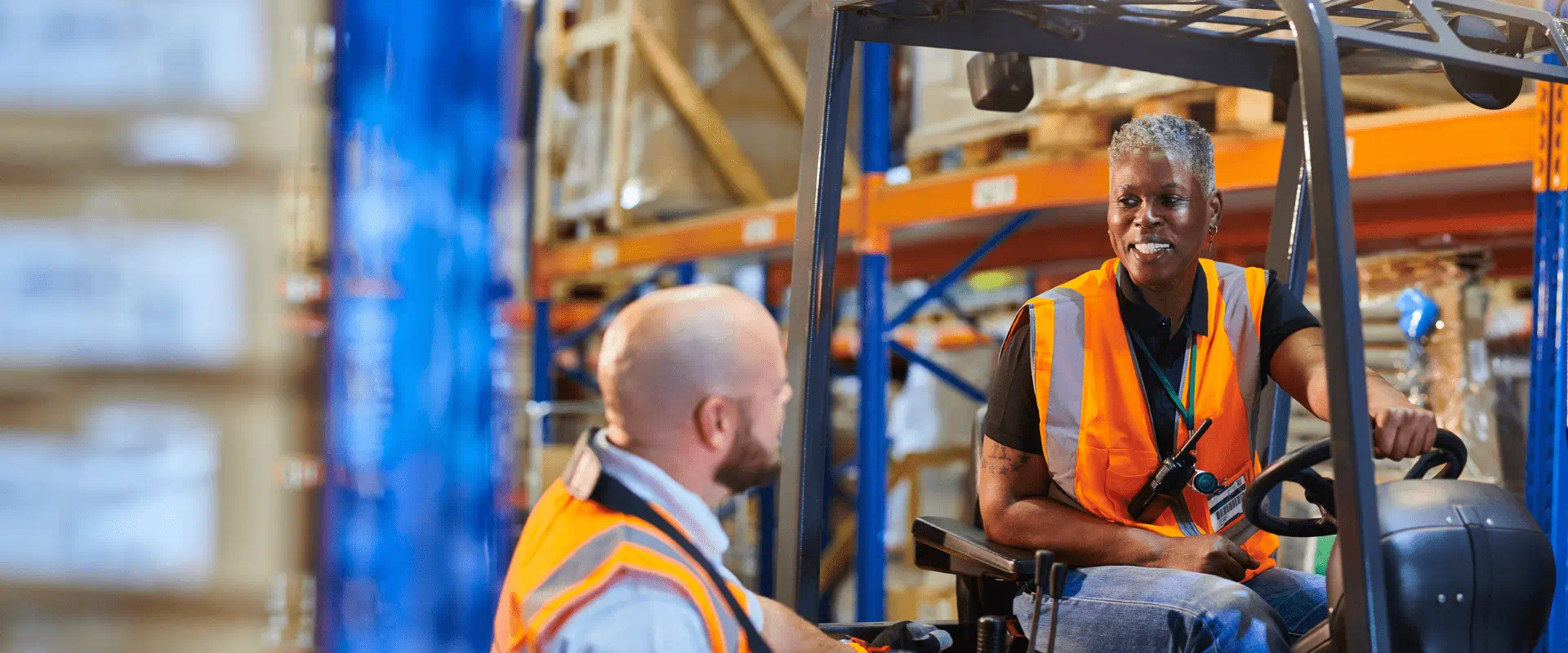 Two workers in safety vests talk in a warehouse. One is operating a forklift. Two workers in safety vests talk in a warehouse. One is operating a forklift.