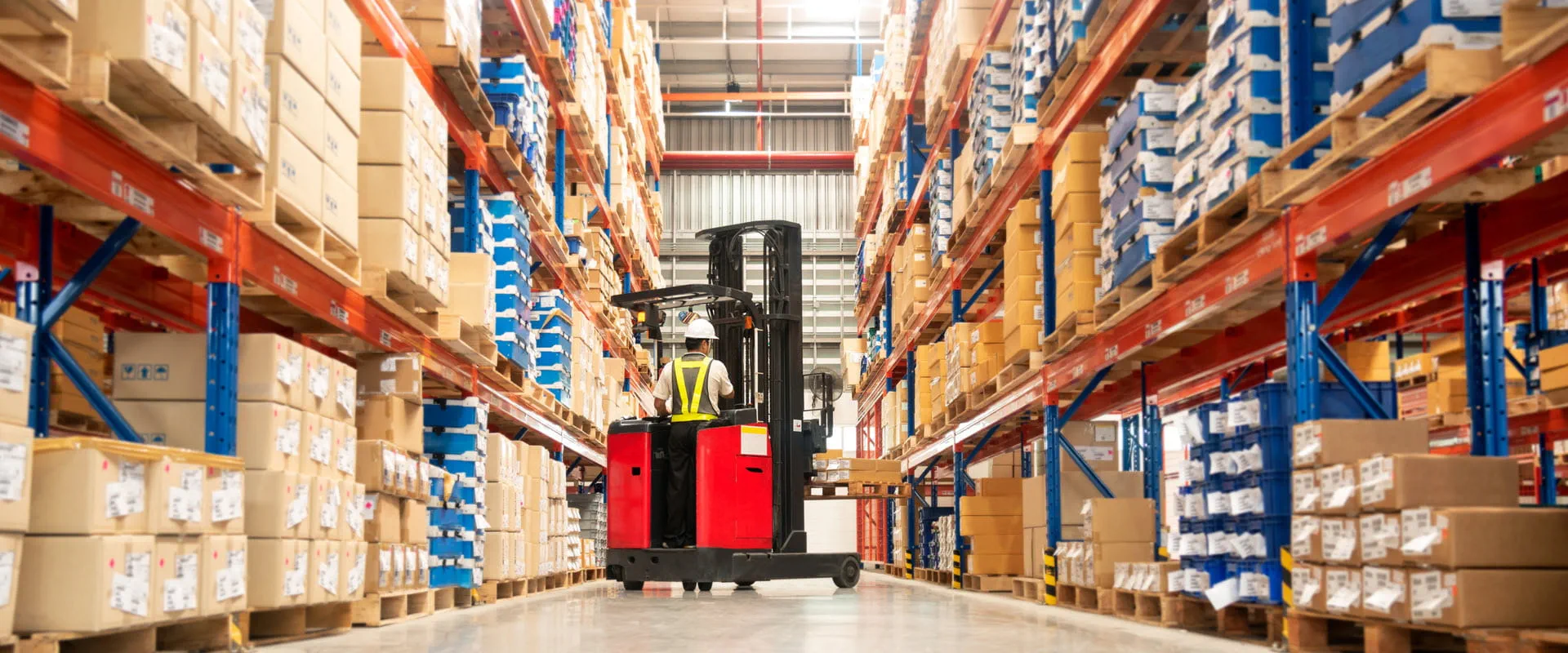 Worker in forklift-truck loading packed goods in huge distribution warehouse with high shelves.