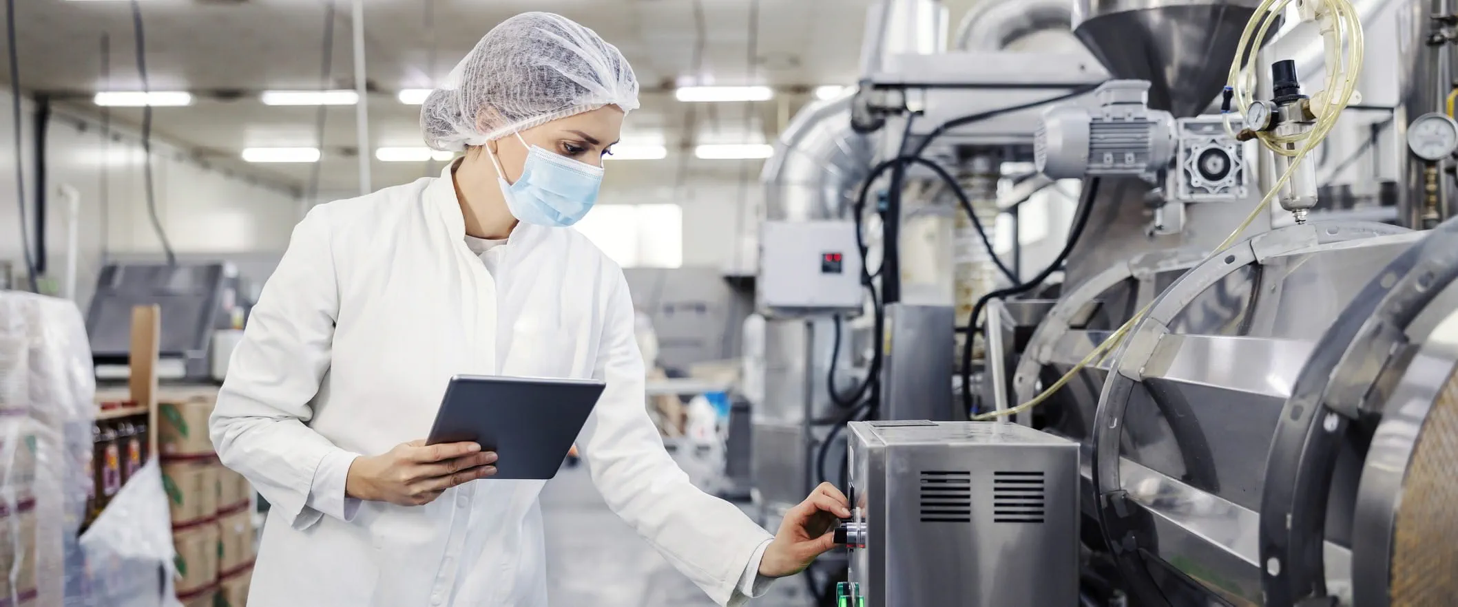 A worker in a hairnet, surgical mask, and white lab coat inspects a piece of machinery. 