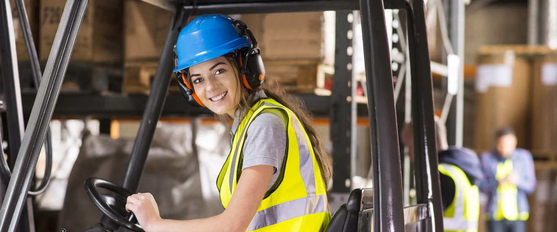 A young female is sitting at the wheel of a forklift truck in a warehouse.