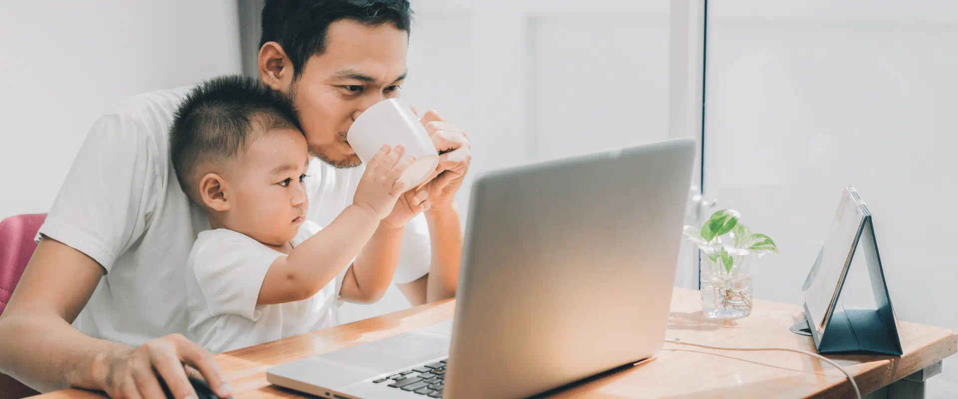 A person sits at home, working on their laptop with a child in their lap, holding their coffee cup.