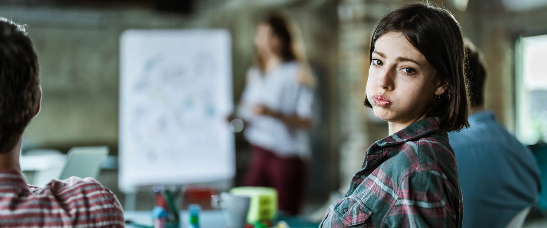A young worker leans back in their chair, sighing and looking exasperated. In the background, their boss is leading a meeting.