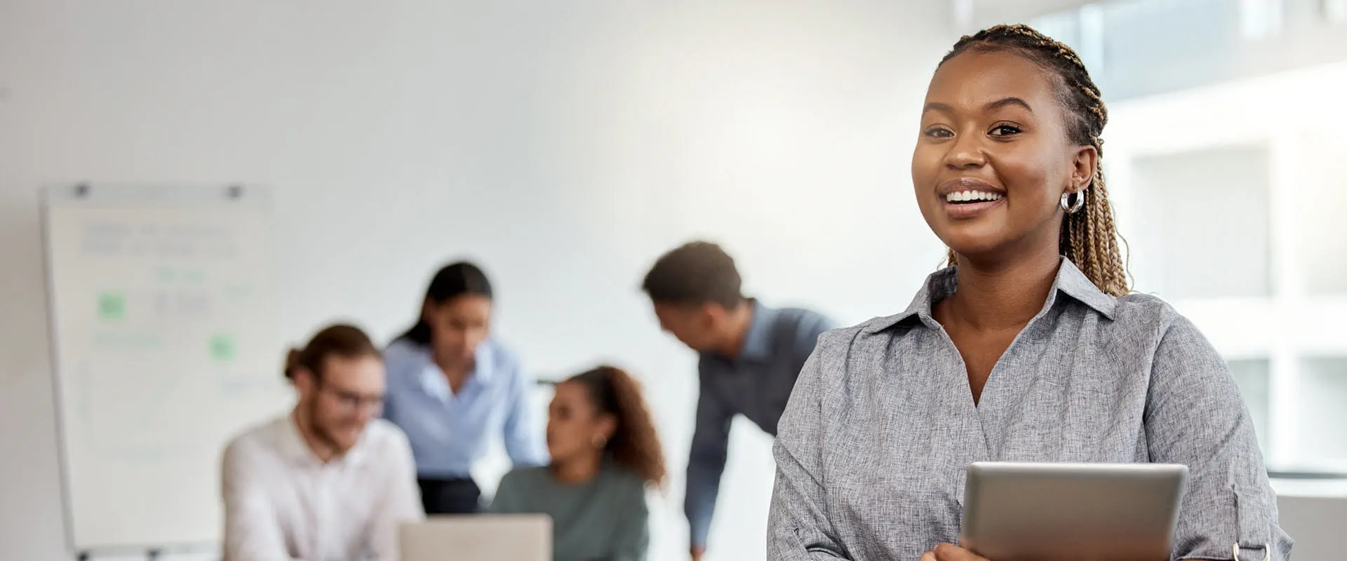 A Black worker holding a tablet stands in front of a conference table where her coworkers are having a meeting.