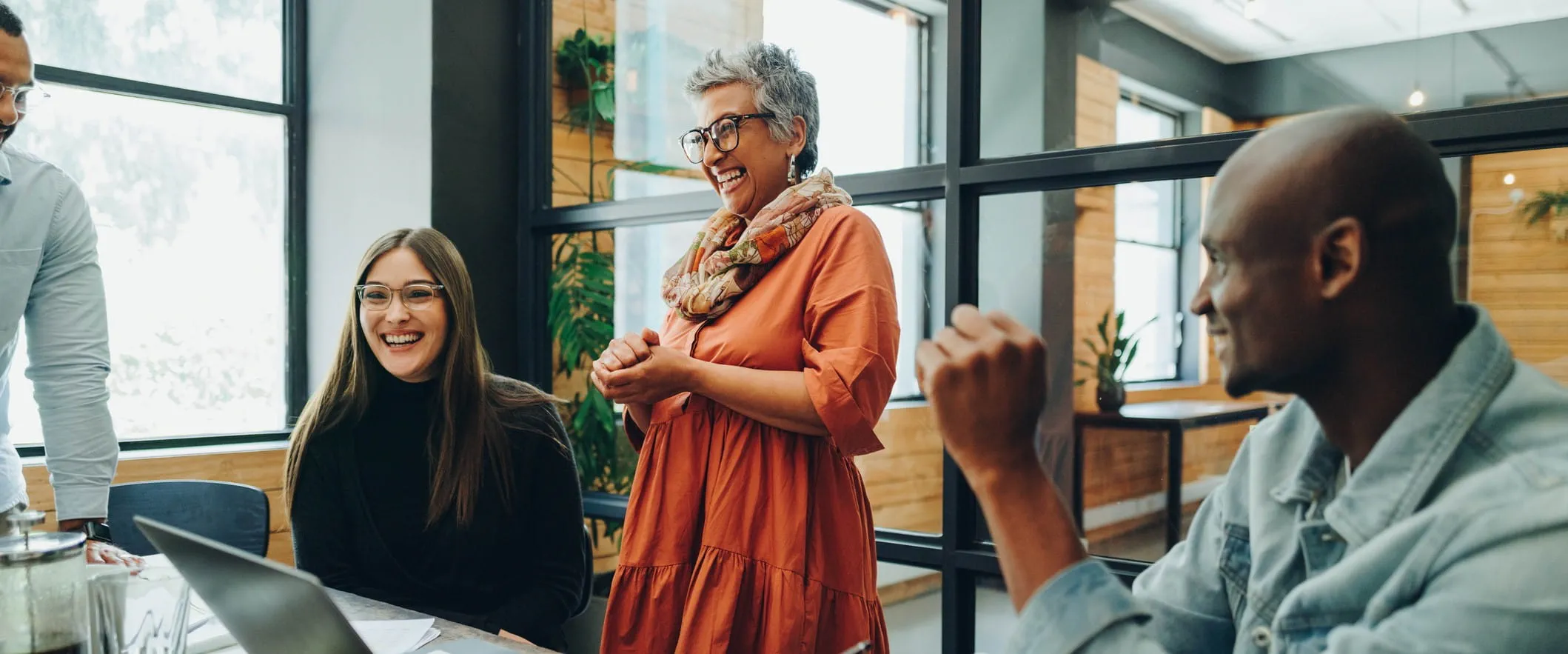 An laughing, older manager stand at an office conference table. Other workers are sitting and standing around the table laughing