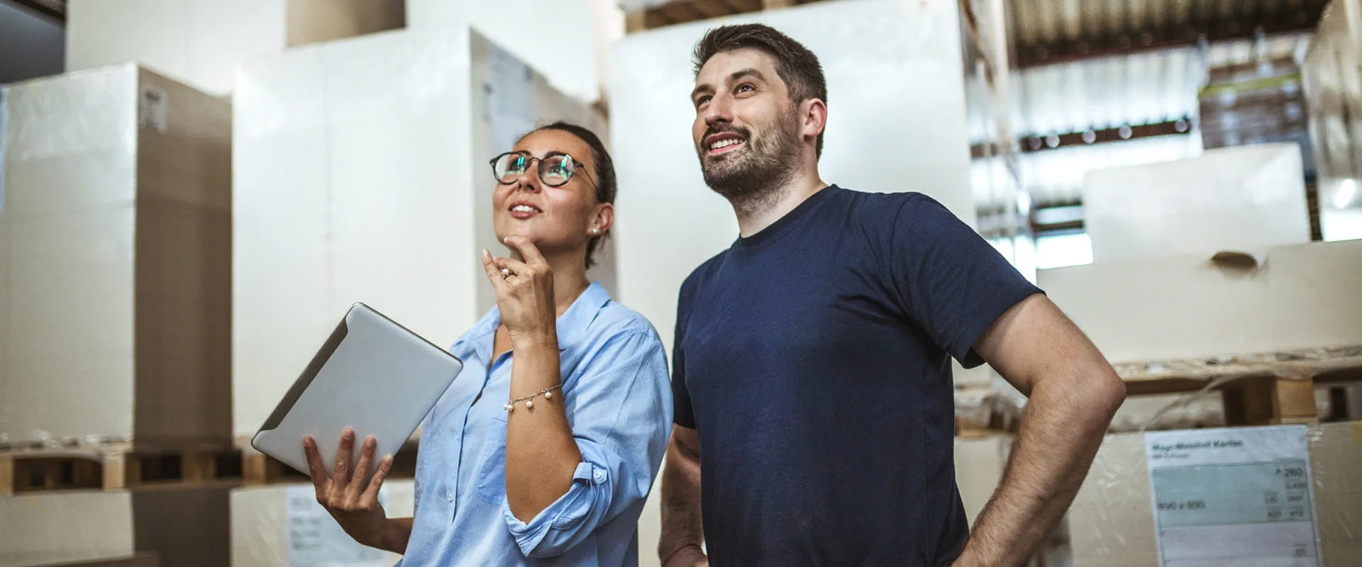 Two workers in a warehouse looking upward. One is holding a tablet.