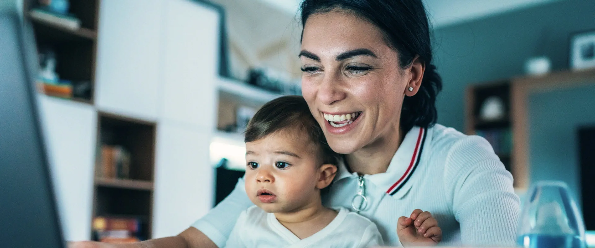 A smiling parent at home, sits at their work laptop with a baby on their lap.