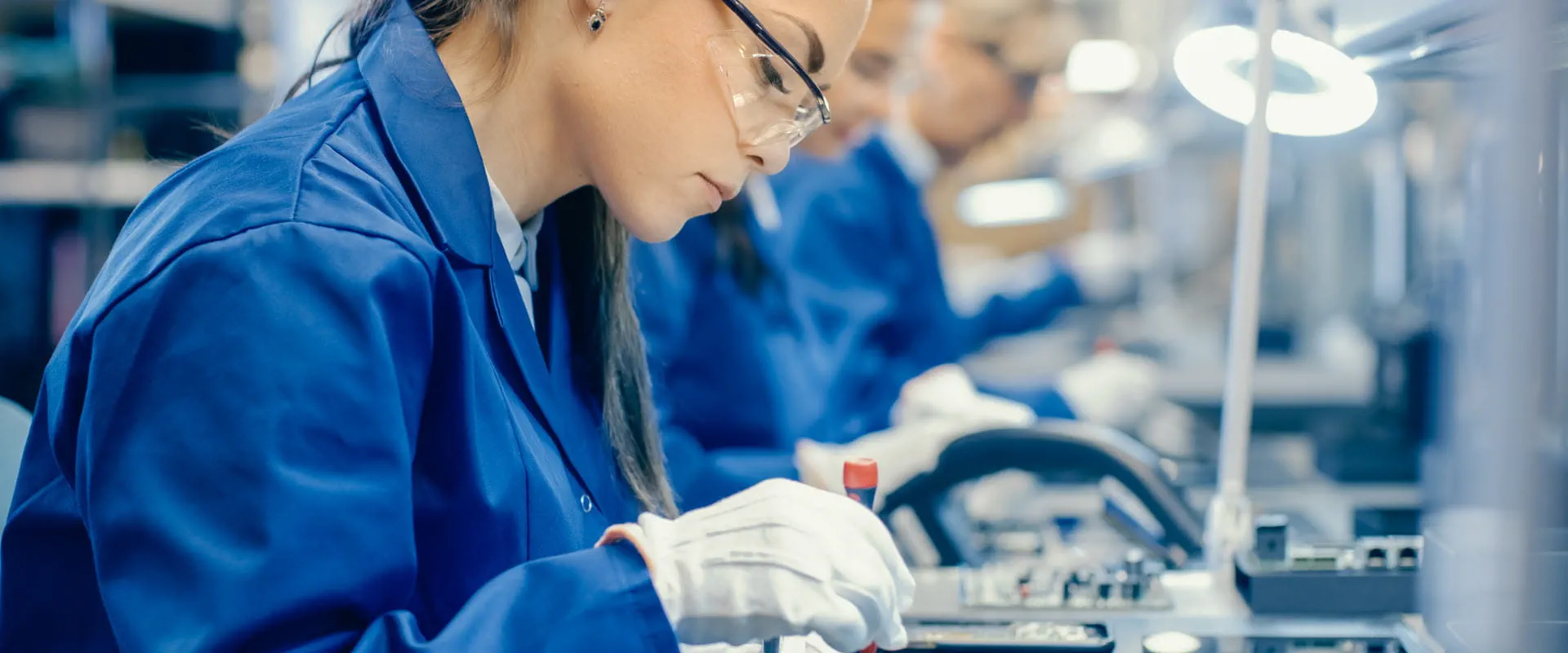 A worker dressed in blue coveralls, sits at a long table, inserting computer chips into devices.