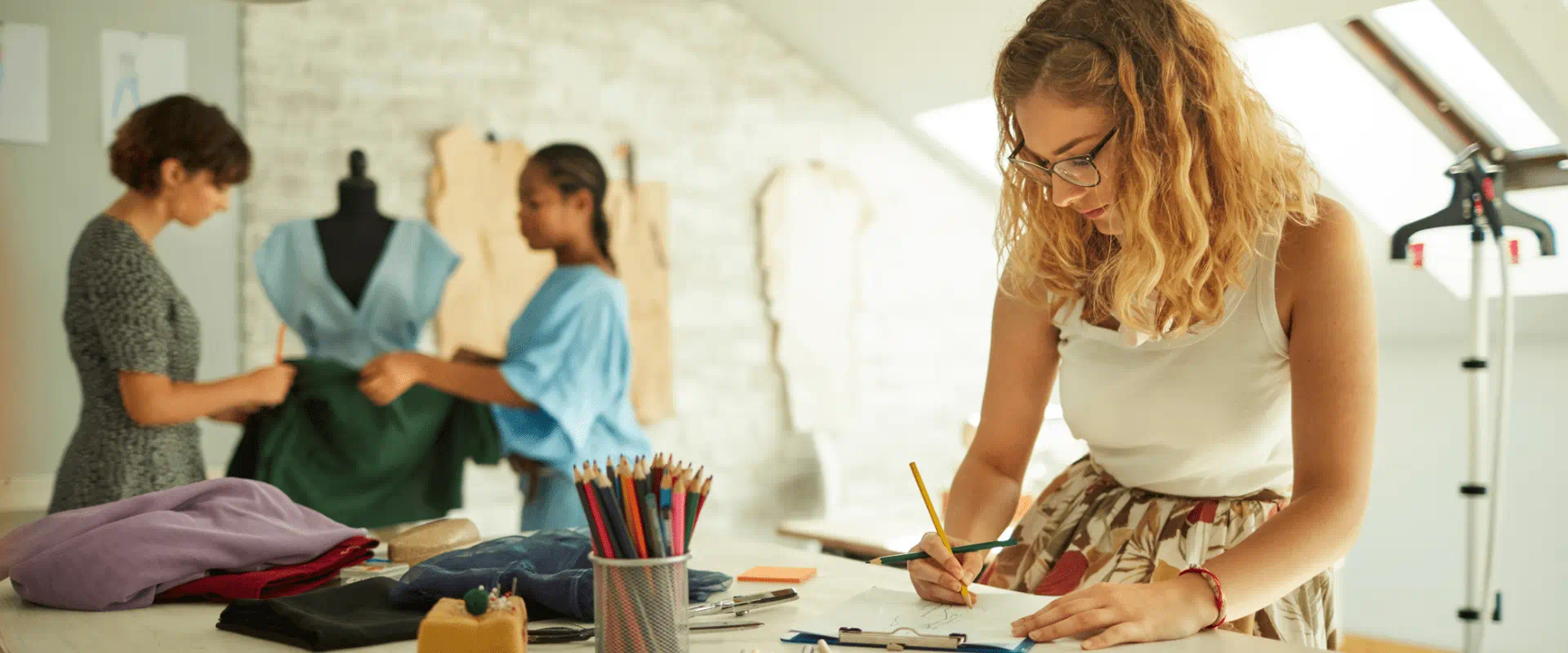 Three interns sit and stand around a worktable, looking at a mannequin or sketching in a notebook