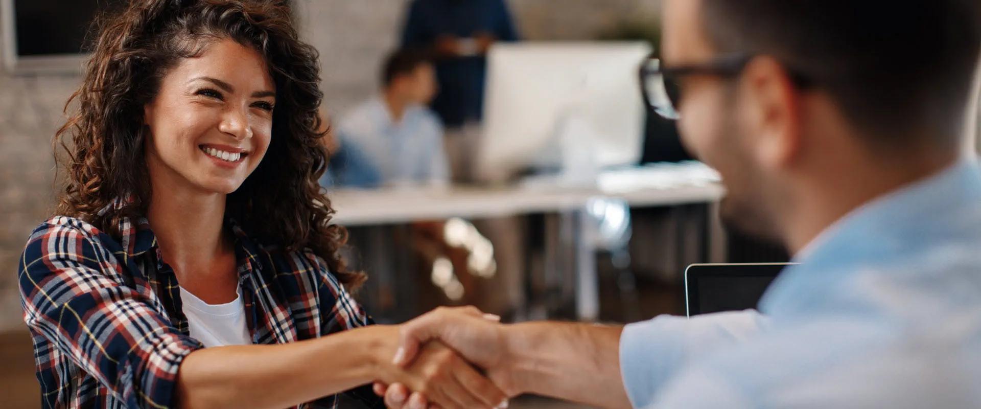 Individual in a plaid shirt shakes hands with another person across the table