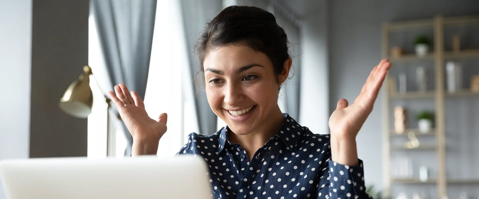 A person wearing a navy blue polka-dot shirt smiles and gestures excitedly while looking at a laptop screen.