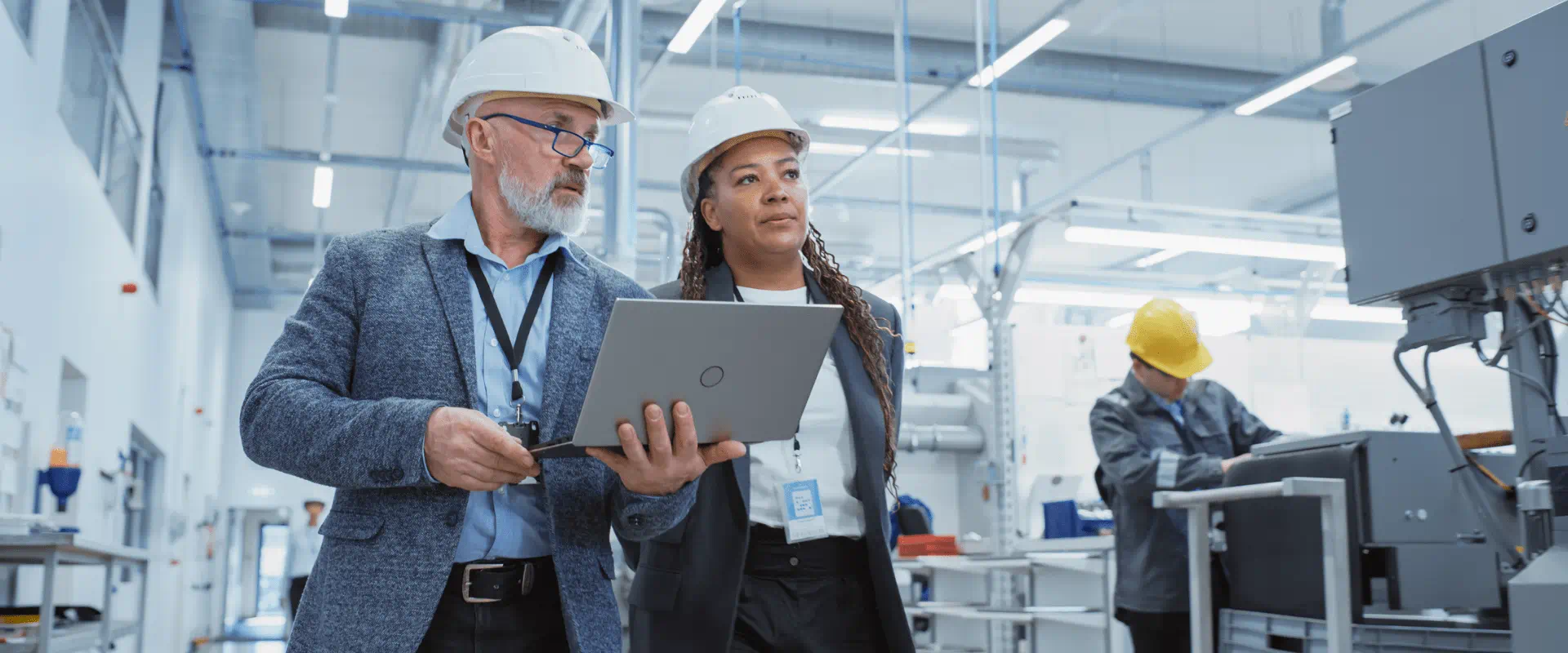 Two people in hard hats, one holding a tablet, walk a factory floor
