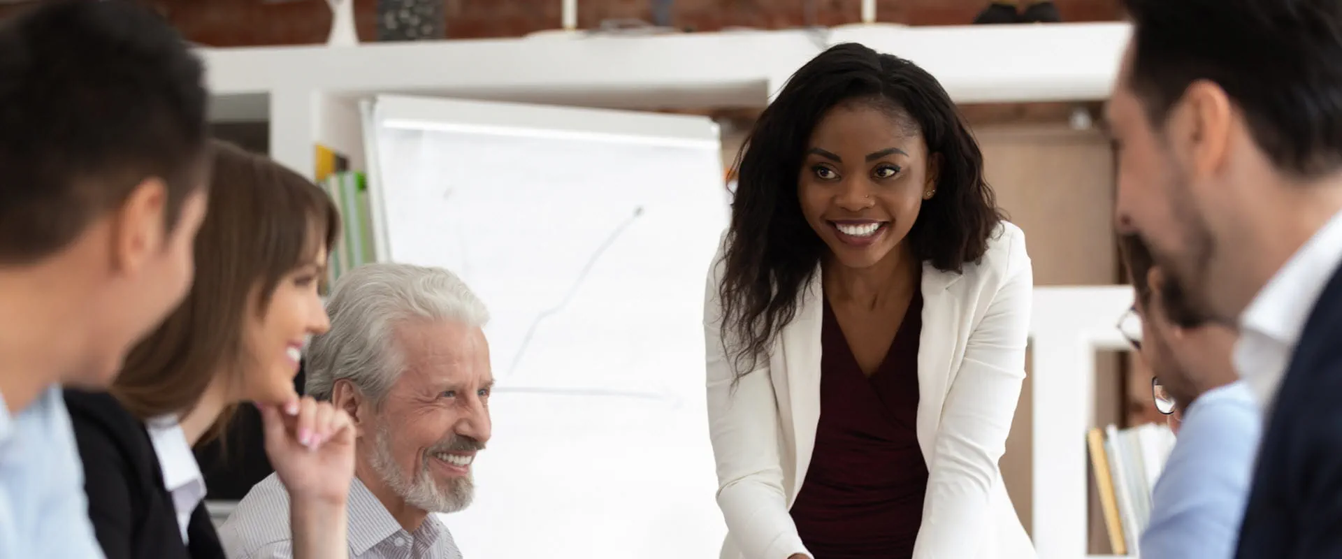 A smiling professional in a white blazer leads a discussion with a diverse group of colleagues in a modern office setting. 