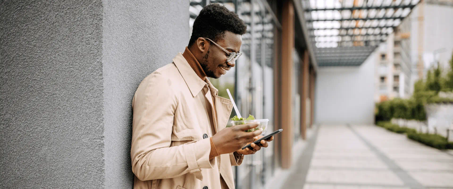 A young person leans against a building, smiling and looking at their phone A young person leans against a building, smiling and looking at their phone