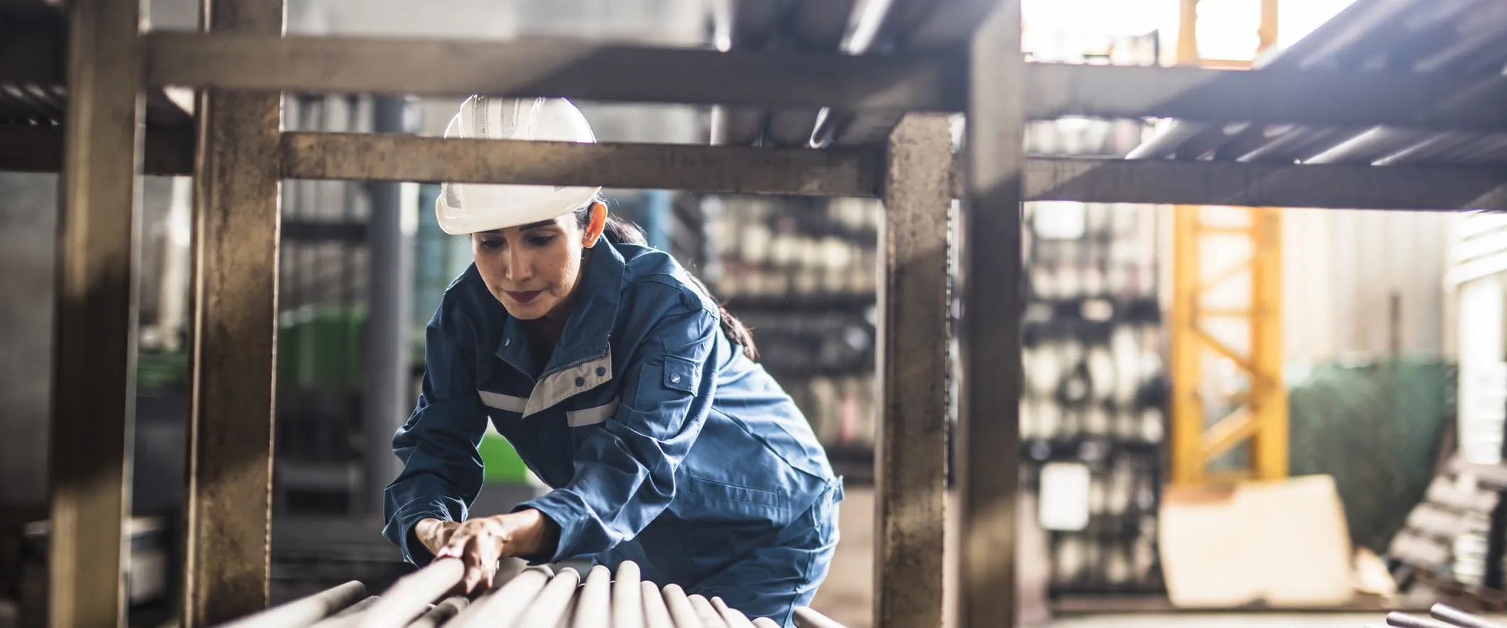 A worker in coveralls and safety gear pulls a long metal tube from a pile of metal tubes.