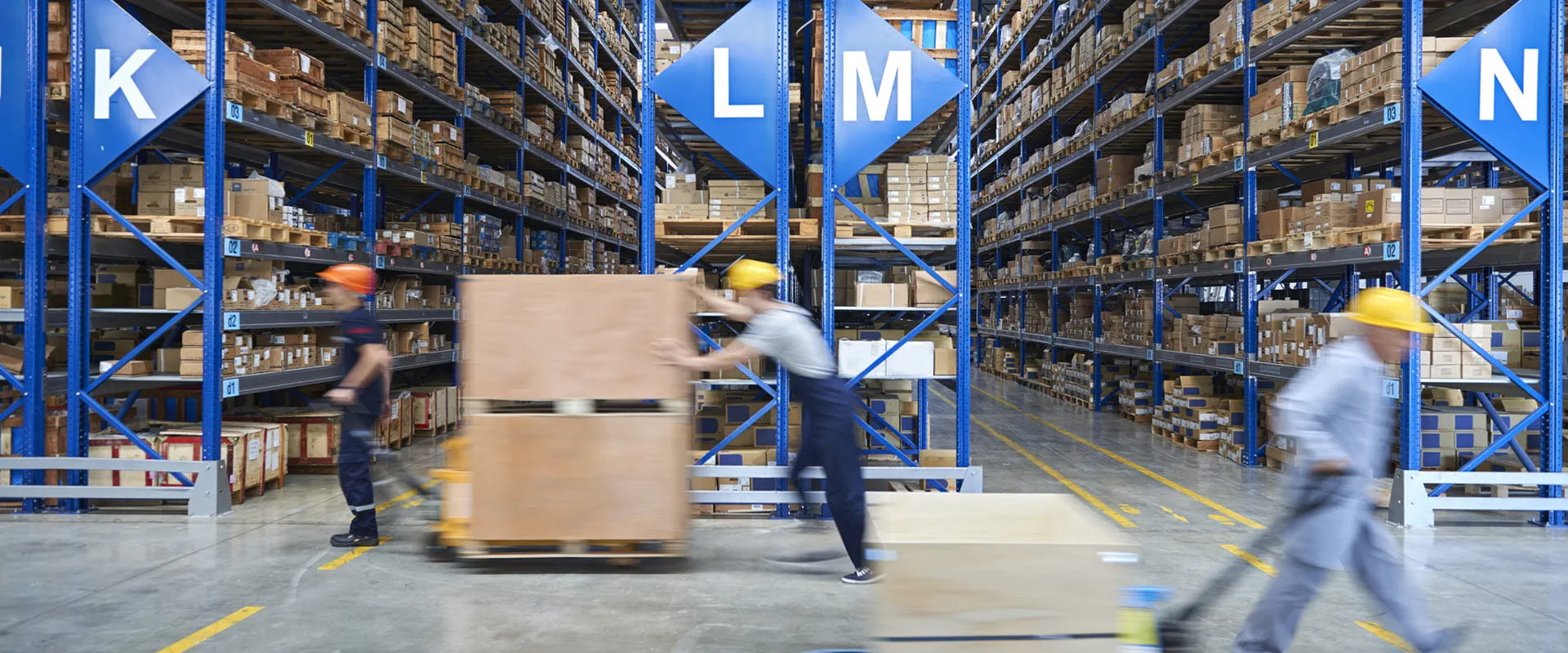Workers briskly walk across the floor of a warehouse filled with floor-to-ceiling shelves, filled with boxes