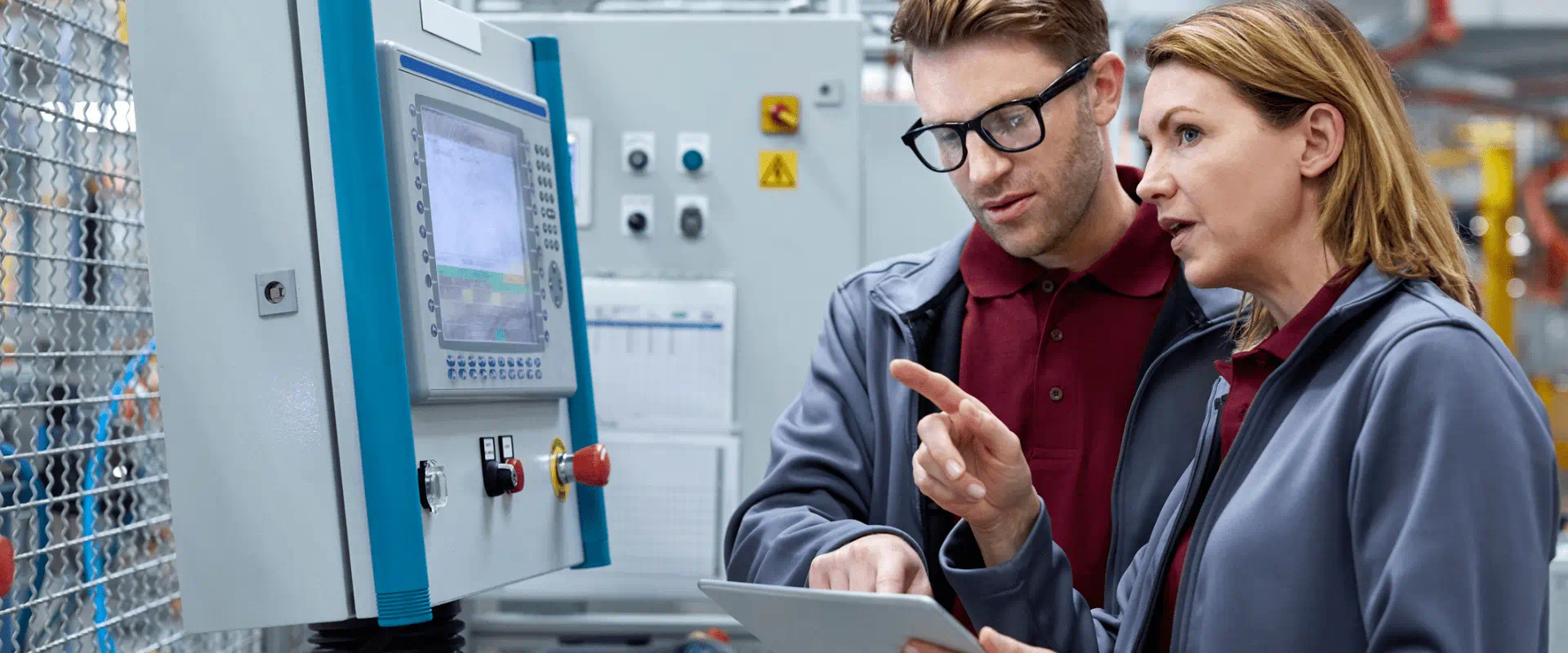 Two workers in an industrial environment, talk while looking at a screen that's attached to a large machine