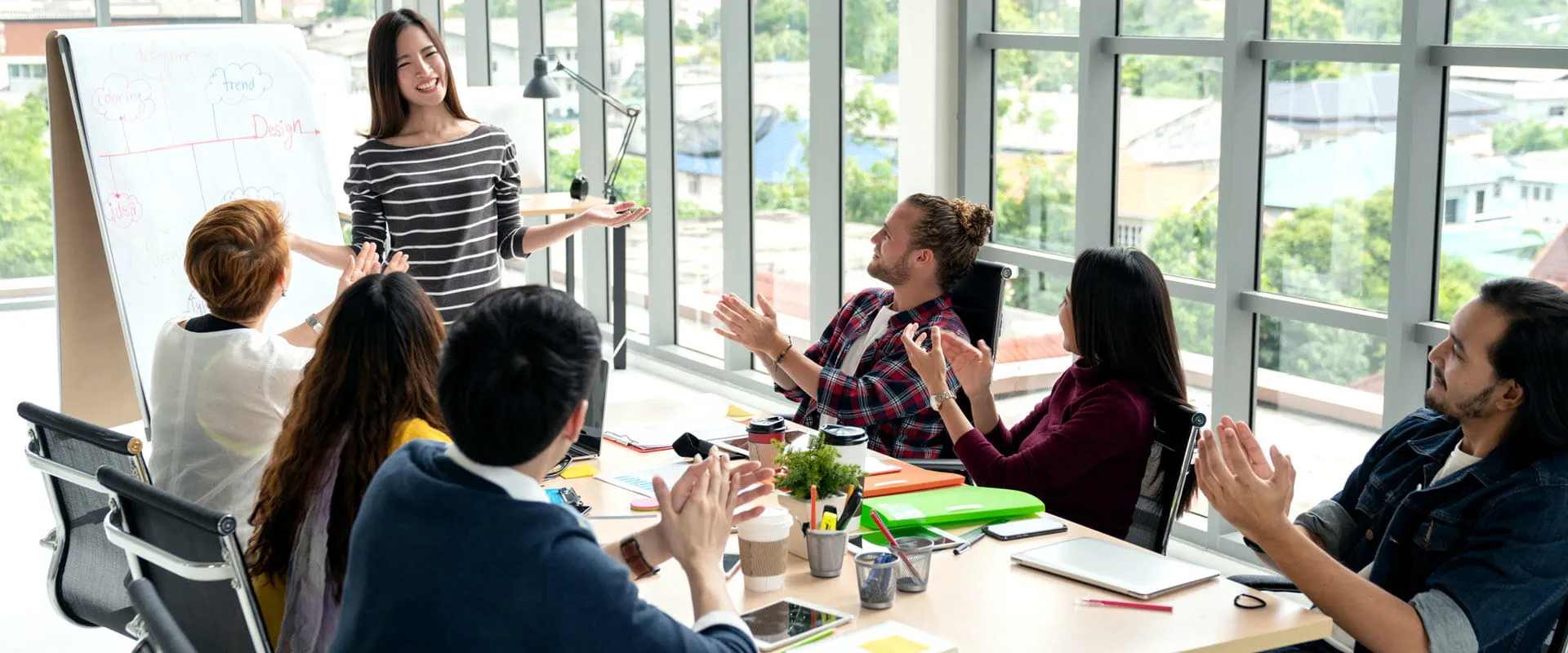 A group of people in a modern office applauding a colleague who is presenting at a whiteboard during a team meeting.