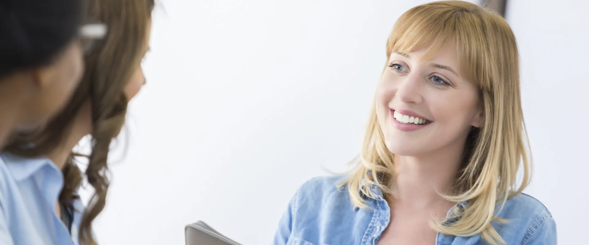 A smiling person in a denim shirt engages in conversation with two colleagues in a bright, professional setting. A smiling person in a denim shirt engages in conversation with two colleagues in a bright, professional setting.